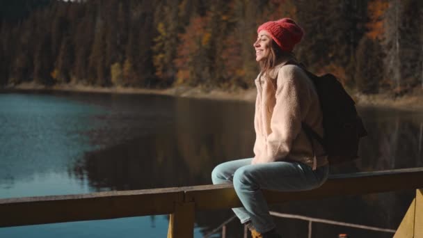 Retrato de inspirada hermosa mujer sonriente excursionista mochilero sentado en el muelle de madera en el hermoso lago de montaña con agua clara de color turquesa y disfrutando de increíble paisaje de otoño tranquilo — Vídeos de Stock