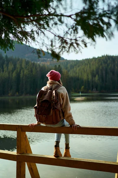 Takanäkymä retkeilijä nainen istuu laiturilla kauniilla vuoristojärvellä ja nauttii metsämaisemasta — kuvapankkivalokuva