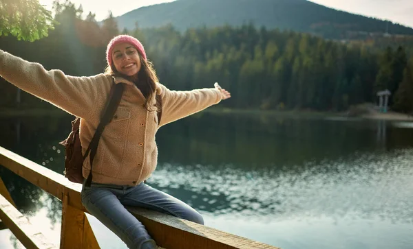 En glad, morsom kvinne sitter på en brygge nær en fjellsjø og nyter naturlandskapet – stockfoto