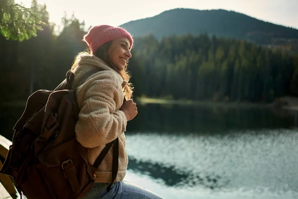 Side View reppumatkailija hymyilevä nainen ihailee vuori järvi keskuudessa metsä — kuvapankkivalokuva