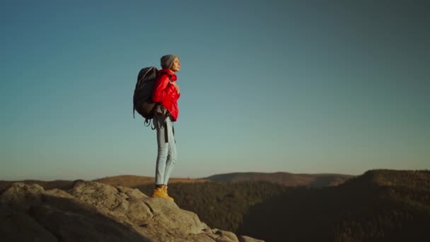 Ralenti vue de côté de la femme randonneur en veste rouge vif avec sac à dos debout sur le bord de la falaise sur fond de coucher de soleil ciel sur les montagnes et regardant vers l'avenir — Video