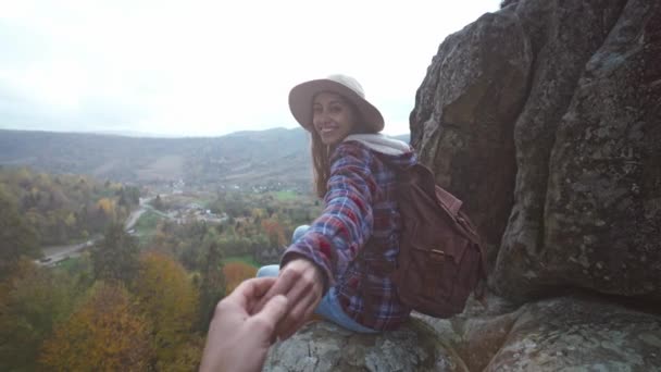Вид от первого лица мужчины, протягивающего руку симпатичной туристке в шляпе с рюкзаком, сидящей на краю скалы на фоне закатного неба над горами — стоковое видео