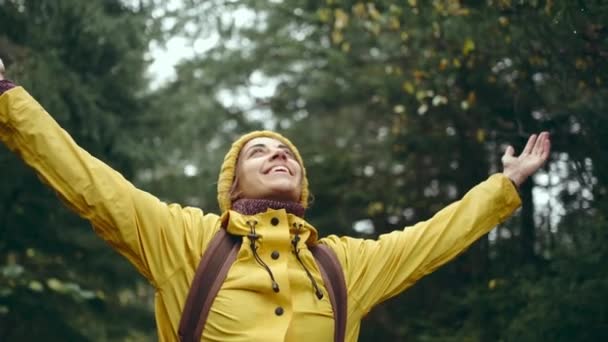 鮮やかな黄色のレインコートとビーニーを着て、バックパックでインスピレーションを受けた若いきれいな女性ハイカーの肖像画は、素晴らしい秋の森の中で自然の美しさ、新鮮さと自由を楽しんで手を上げて見上げ — ストック動画