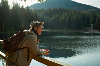 Sırt çantalı bir adam dağ gölüne hayran, ormandaki temiz havanın tadını çıkarıyor.