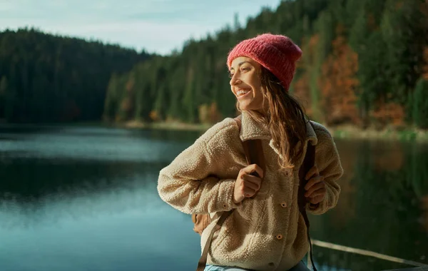 En lystig vandringskvinne som ser bort og smiler, går nær vakker innsjø med skog – stockfoto
