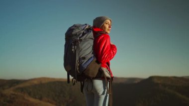 Sırt çantalı, parlak kırmızı ceketli kadın yürüyüşçünün yavaş çekimde arka planda duran günbatımı manzarası dağların üzerinden gökyüzüne bakıyor.