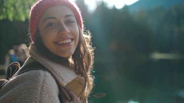 Vista lateral mochileiro mulher sorrindo admirando lago de montanha entre a floresta com luz da manhã ao redor — Vídeo de Stock