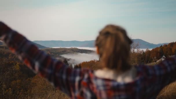 Vista traseira mulher caminhante mochileiro vestindo uma jaqueta quadriculada e caminhadas levantando os braços para o céu. menina apreciando a vista acima das nuvens no topo da montanha no nascer do sol de outono — Vídeo de Stock