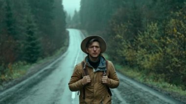 Otostopçu adam eski moda giyiniyor. Sırt çantalı gezgin soğuk ve nemli bir sonbahar gününde alog yolunda yürüyor ve bir araba bekliyor. Adam kameraya bakıyor.