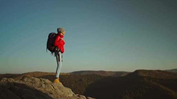 Сзади на вершине горы на закате стоит женщина-туристка в красной куртке с рюкзаком. свобода и одиночные путешествия концепции. единство с природой — стоковое видео