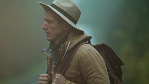 Close up retrato de homem carona vestindo em estilo retro. viajante em chapéu com mochila fica ao ar livre na estrada no frio molhado nebuloso dia de outono e espera um carro — Vídeo de Stock