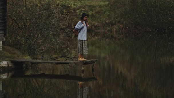 Elegante donna viaggiatore ispirato in cappello con zaino in piedi sul lungomare di legno al lago, guardando la vista della natura autunnale, alberi con brillante riflessione fogliame arancione in acqua — Video Stock