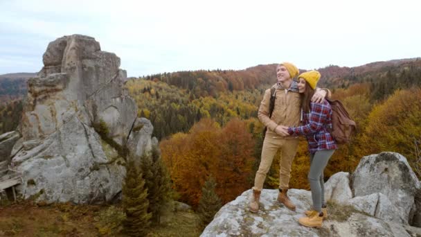 Пара улыбающихся туристов стоит на фоне осеннего пейзажа со скалами в национальном парке Тустань, Украина. любящий нежный мужчина и женщина, путешествующие вместе и единение с природой — стоковое видео