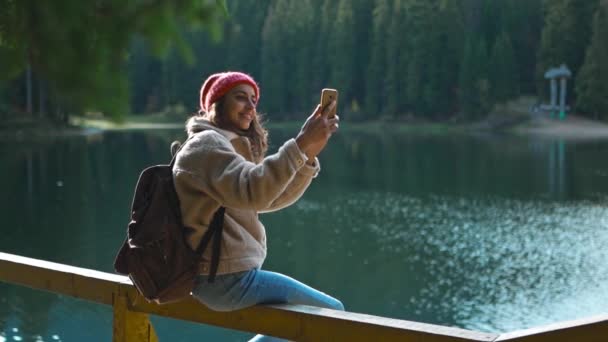Venti Hiker Fotografo Hipster donna nel bosco riprese Selfie Lake View. Travel Girl Videografa stand in autunno Natura Scenico e riprese di se stessa sulla fotocamera dello smartphone, fare videochiamate — Video Stock