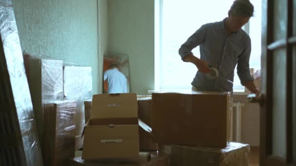 Millennial männliche Mieter im Wohnzimmer mit Klebeband Spender Versiegelung Kartons Umzug in ein neues Haus. Mietwohnung verlassen, Umzugstag und persönliche Sachen packen — Stockvideo