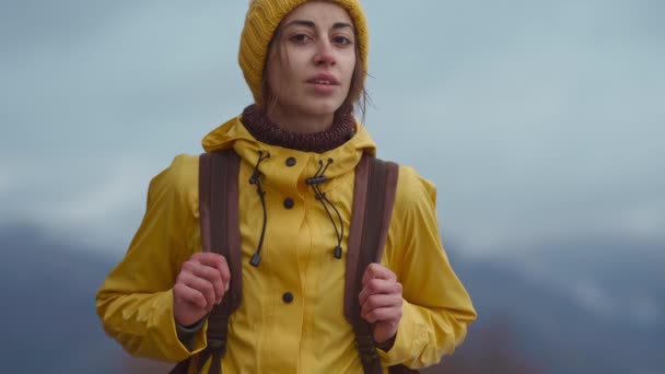 Primer plano retrato de mujer atractiva excursionista en impermeable de color amarillo brillante y gorro, con la mochila se encuentra al aire libre en el frío día nublado de otoño y mirando a la cámara y sonriendo — Vídeos de Stock