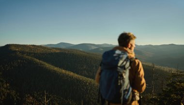 Gün batımında dağların ve tepelerin panoramik manzarası, insan yürüyüşçüsünün odaklanmamış silueti.
