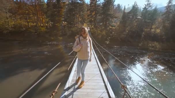 Rallentatore ritratto di felice bella giovane donna turista in piedi su ponte sospeso in legno appeso sul fiume di montagna. paesaggio incredibile con abeti rossi, montagne e cielo sullo sfondo — Video Stock