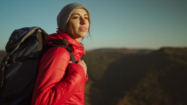Slow motion portrait woman hiker in bright red jacket with backpack stands on mountain top against background of sunset sky over mountains, looking ahead and enjoying freedom and successful ascent — 图库视频影像