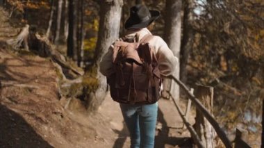 Arkaya bakan kadın turist, güneşli bir sonbahar gününde sonbahar ormanında yolda yürüyor. Sırt çantasıyla dağ gölünün etrafındaki ormanda yürüyüş yapan kadın yürüyüşçü. Soğuk güneşli bir sabahta, açık hava macerası.