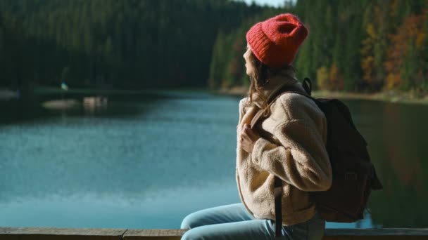 Terug uitzicht geïnspireerde vrolijke vrouw wandelaar zit op houten pier bij prachtige bergmeer met turquoise helder water en geniet van verbazingwekkende rustige herfst landschap. wilde natuur schoonheid, gezonde actieve levensstijl — Stockvideo