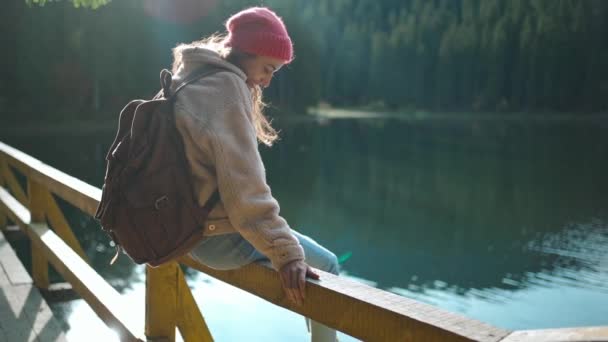 Câmera lenta inspirada mulher alegre caminhante senta-se no cais de madeira em belo lago de montanha com água azul-turquesa e goza de incrível paisagem calma outono. natureza selvagem beleza, estilo de vida ativo saudável — Vídeo de Stock