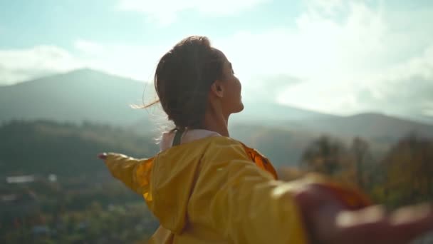 Молодая женщина в жёлтой куртке поднимает руки в голубое небо и наслаждается видом над облаками на вершине горы — стоковое видео