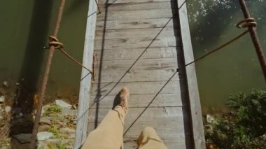 Hipster bir adamın yavaş çekim görüntüsü dağ nehri üzerinde asılı duran ahşap bir köprüde yürüyor. Erkek bacaklarının günlük seyahat çizmeleriyle üst görüntüsü