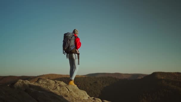Αργή κίνηση άποψη από πίσω περιπετειώδη γυναίκα πεζοπόρος backpacker σε κόκκινο σακάκι με σακίδιο στέκεται στην κορυφή του βουνού στο ηλιοβασίλεμα πάνω από το δάσος. ελευθερία και σόλο ταξιδιωτική έννοια. ενότητα με τη φύση — Αρχείο Βίντεο