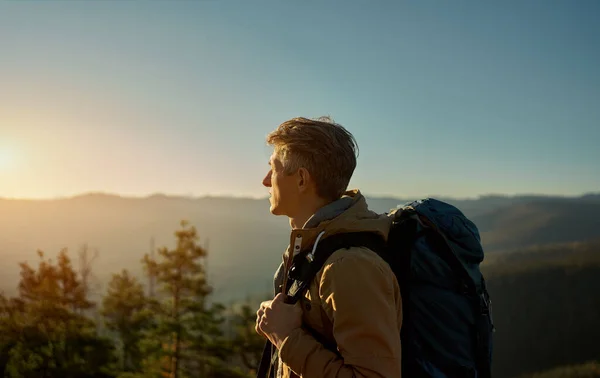 Mies matkaaja reppu seisoo auringonlaskussa valossa vuorten ja kukkuloiden — kuvapankkivalokuva