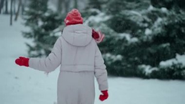 Kamera, mutlu ve heyecanlı kadını kar yağarken parkta eğlenceli bir şekilde koşarken takip ediyor. Ağaçlar soğuk kış gününde karın üzerinde duruyor.