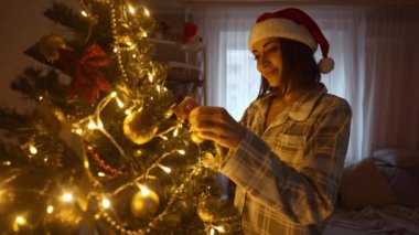 Noel şapkalı mutlu kadının yavaş çekimde çekilmiş portresi Noel ağacının dalına altın topu asıyor. Noel 'i ve yeni yılı evde kutlamaya hazırlanıyorum.