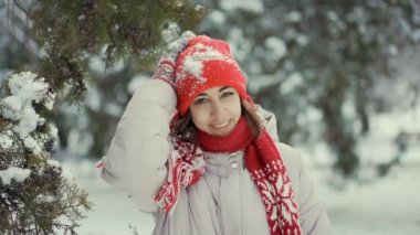 4k ağır çekim mutlu gülümseyen kadın örgü kırmızı bereli, atkı ve eldivenli. Karla kaplı şapkalı kız karlı köknar ağacının orada duruyor ve kameraya bakıyor. Kışın dışarıda eğleniyoruz.