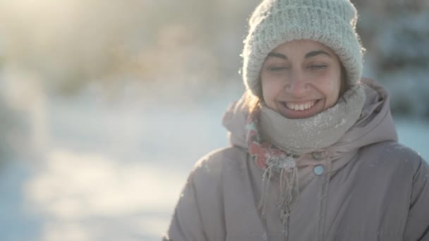 4k slow motion close up face of beautiful smiling woman in knitted beanie and scarf standing in park at winter outdoors. snow is on girl cap, face and eyelashes. woman cheerfuly smiles — Stock Video
