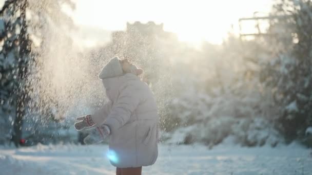 Playful young woman in warm clothes throws snow up, circles around her and clapping hands with snowflakes in sunny winter snowy park. — Stock Video