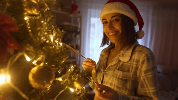 산타햇에 있는 행복 한 여인의 느린 동작 초상화가 크리스마스 트리의 가지에 황금 공을 걸어 놓는다. 집 장식 과 크리스마스 및 새해 맞이를 준비하는 모습 — 비디오