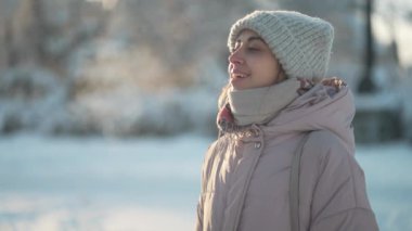 Kış parkında, kıvırcık güneşli bir günde örgü şapkalı ve atkılı mutlu bir kadın. Kadın gün ışığında buhar üflüyor. Kış tatilin kutlu olsun, iyi eğlenceler..