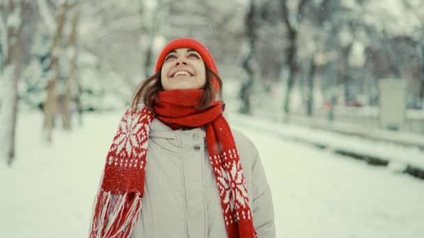 Счастливое зимнее время, радость для первого снега. 4k портрет замедленной съемки красивая улыбающаяся молодая женщина в парке, вязаная красная шапочка и варежки, стоящие в снежном парке, глядя вверх и приветствуя снежинки — стоковое видео