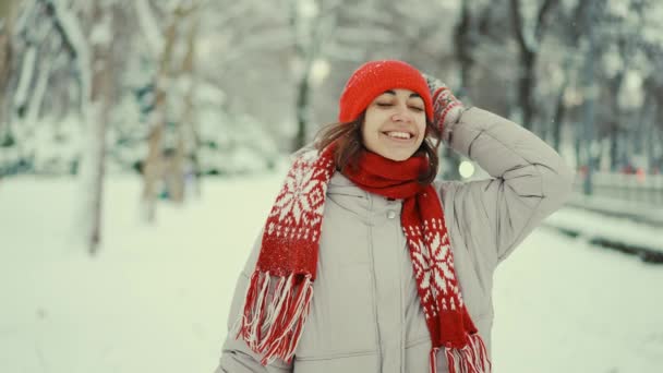 4k slow motion cheerful woman in red cap and mittens enjoying first snow in winter park in january. portrait attractive smiling girl in winter wear looking on snowflakes around with delight — Wideo stockowe