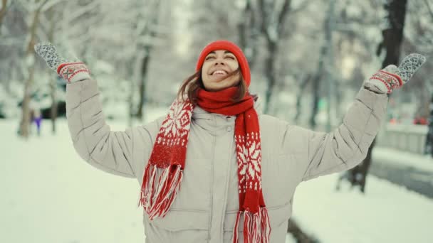 Cheerful smiling woman in warm clothes, red knitted cap, scarf and mittens stands in snowy park enjoying raising arms and catching snowflakes after blizzard in city. Happy woman playing with snow – Stock-video