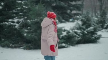 Kamera, mutlu ve heyecanlı kadını kar yağarken parkta eğlenceli bir şekilde koşarken takip ediyor. Ağaçlar soğuk kış gününde karın üzerinde duruyor.