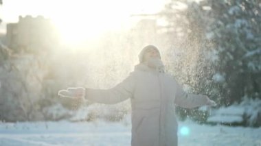 Sıcak giysiler içindeki neşeli genç kadın karları fırlatıyor, etrafında daireler çiziyor ve güneşli kış parkında kar taneleriyle el çırpıyor..