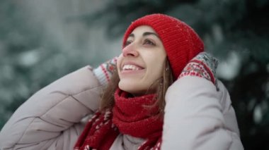 close up slow motion candid smiling happy woman in red woolen mittens and beanie stands in park in snow, looks and enjoys falling snowflakes