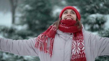beautiful smiling woman in red beanie and scarf standing in forest at snowy winter day outdoors. girl raises hands and looks up on blowing snowflakes