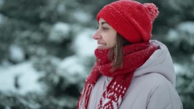 slow motion beautiful smiling woman in red beanie and scarf standing in forest at snowy winter day outdoors. girl turns around and looks on blowing snowflakes