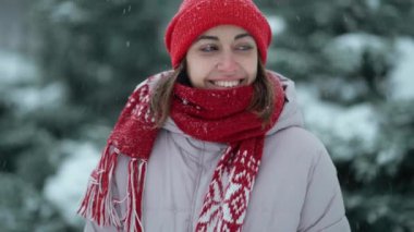 slow motion beautiful smiling woman in red beanie and scarf standing in forest at snowy winter day outdoors. girl turns around, raises hands and looks up on blowing snowflakes