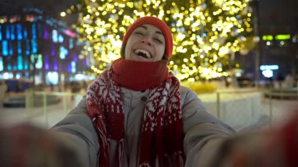Feliz jovem alegre em boné vermelho tomando selfie no meio da rua em luzes do mercado de Natal contra o fundo da árvore de Natal público. ano novo e inverno feriados conceito — Vídeo de Stock