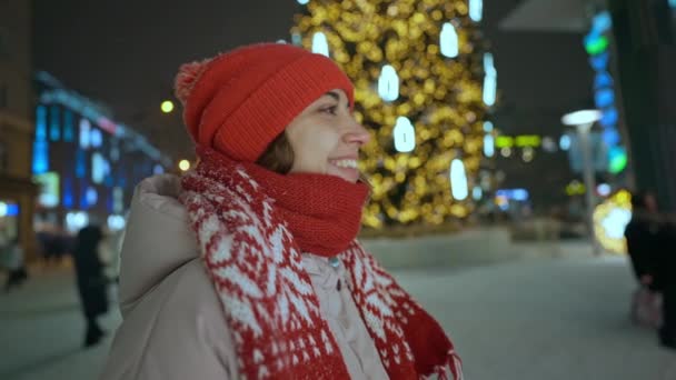 Close up retrato de feliz alegre jovem em boné vermelho e cachecol em pé na praça da cidade à noite contra árvore de natal pública com luzes de fundo. ano novo e inverno feriados conceito — Vídeo de Stock
