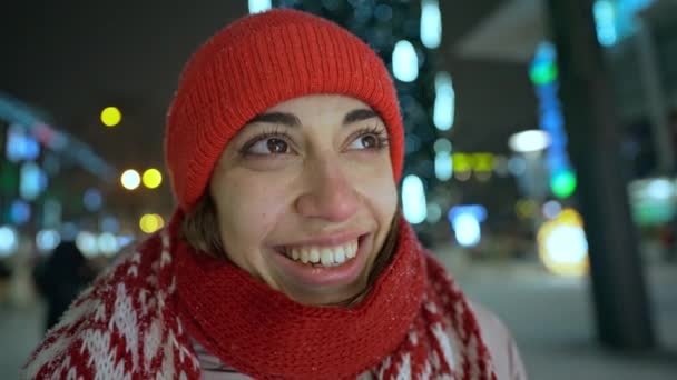 Extreme closeup portrait of smiling cheerful young woman in knitted red cap and scarf standing on city square at night against public christmas tree with lights background. focus on womans eyes — Video