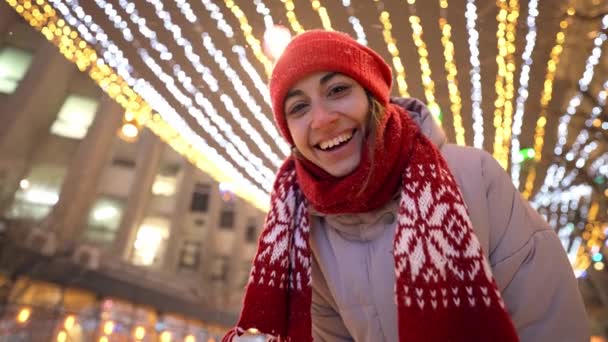 Mujer sonriente feliz en punto gorra roja y bufanda tomar selfie, mirando a la cámara y agitando las manos en la plaza de la ciudad con iluminaciones festivas por la noche. nieve está cayendo y copos de nieve volando alrededor — Vídeos de Stock